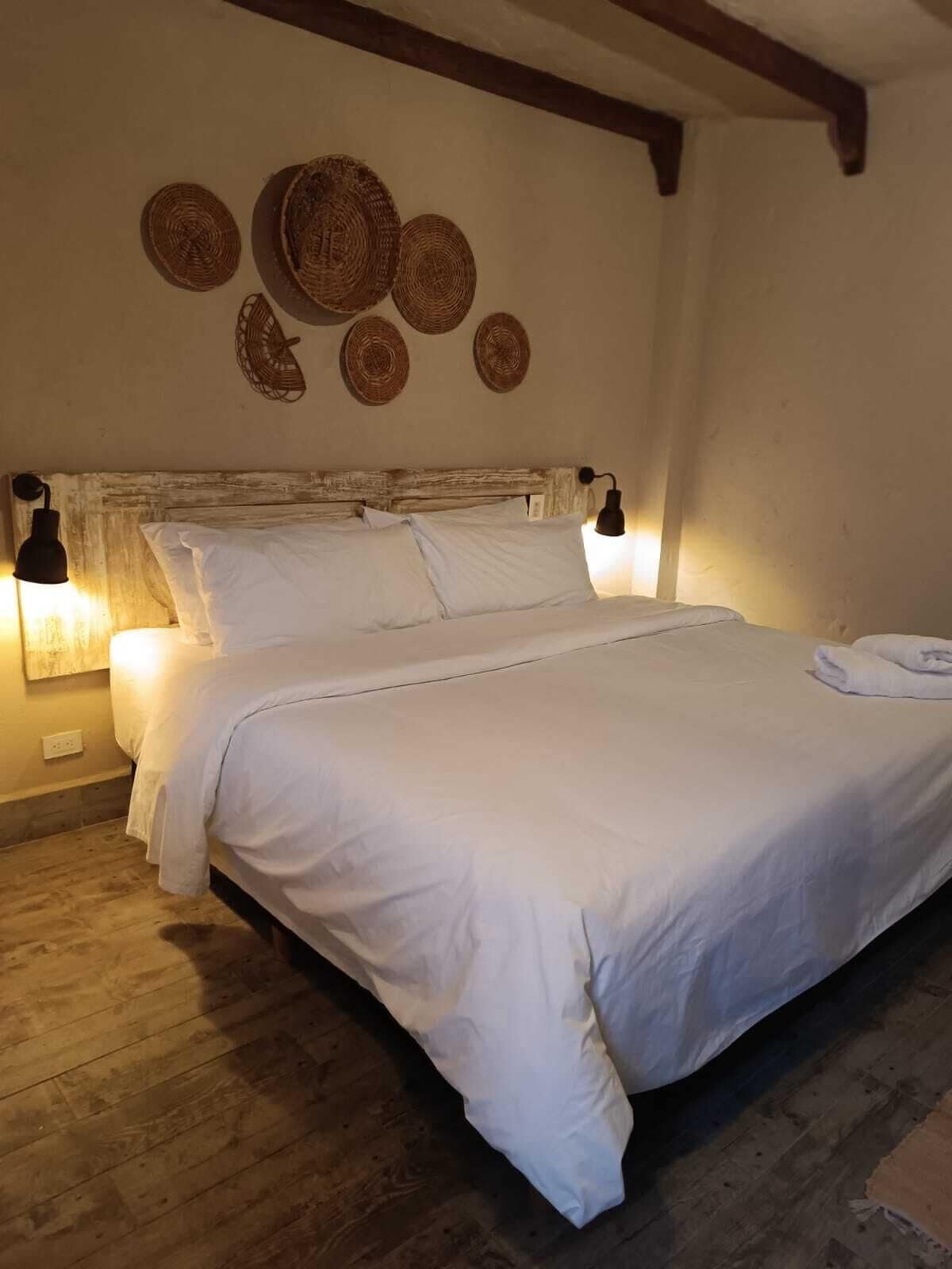 Ein ordentlich bezogenes Bett mit weißen Textilien, zwei Wandleuchten und Wandbehängen aus Gewebe.