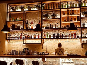 Un bar avec des étagères remplies de bouteilles et de verres, un barman et un mur en pierre rustique.