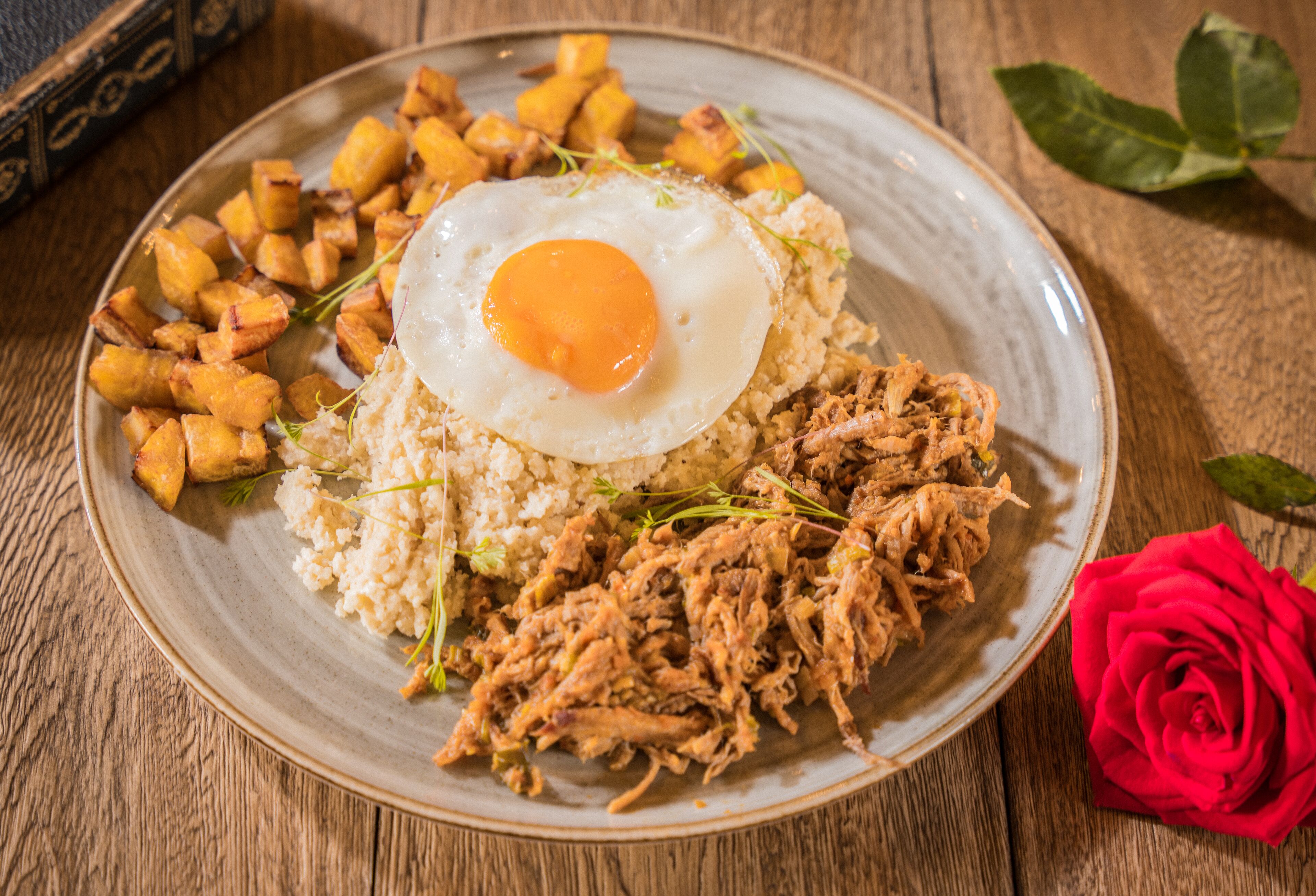 Un plato de comida con un huevo frito, carne desmenuzada y patatas asadas sobre una mesa de madera.