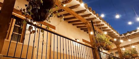 Un espacio al aire libre techado con vigas de madera, barandilla metálica y plantas colgantes.