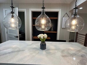 Une table à manger en marbre, ornée d’un vase de fleurs blanches, trois lampes suspendues et une porte visible au fond.