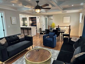 Un salon moderne avec un ventilateur de plafond, des canapés noirs, une table à manger et un coin cuisine à l’arrière-plan.