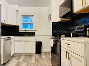 A modern kitchen with white cabinetry, a black countertop, a stainless steel oven, and a black trash bin.