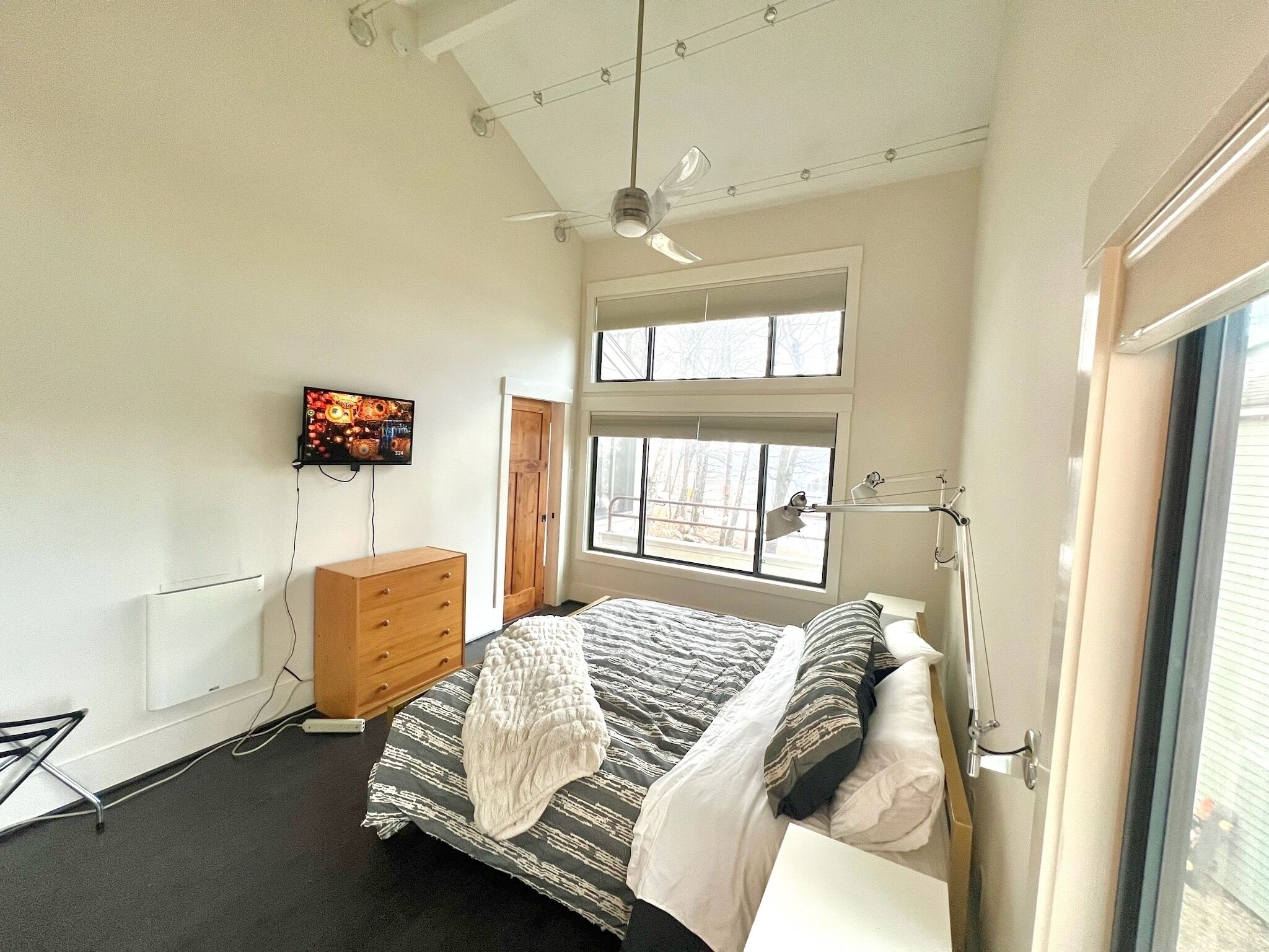 A bedroom with a bed, a TV mounted on the wall, a dresser, and a window.
