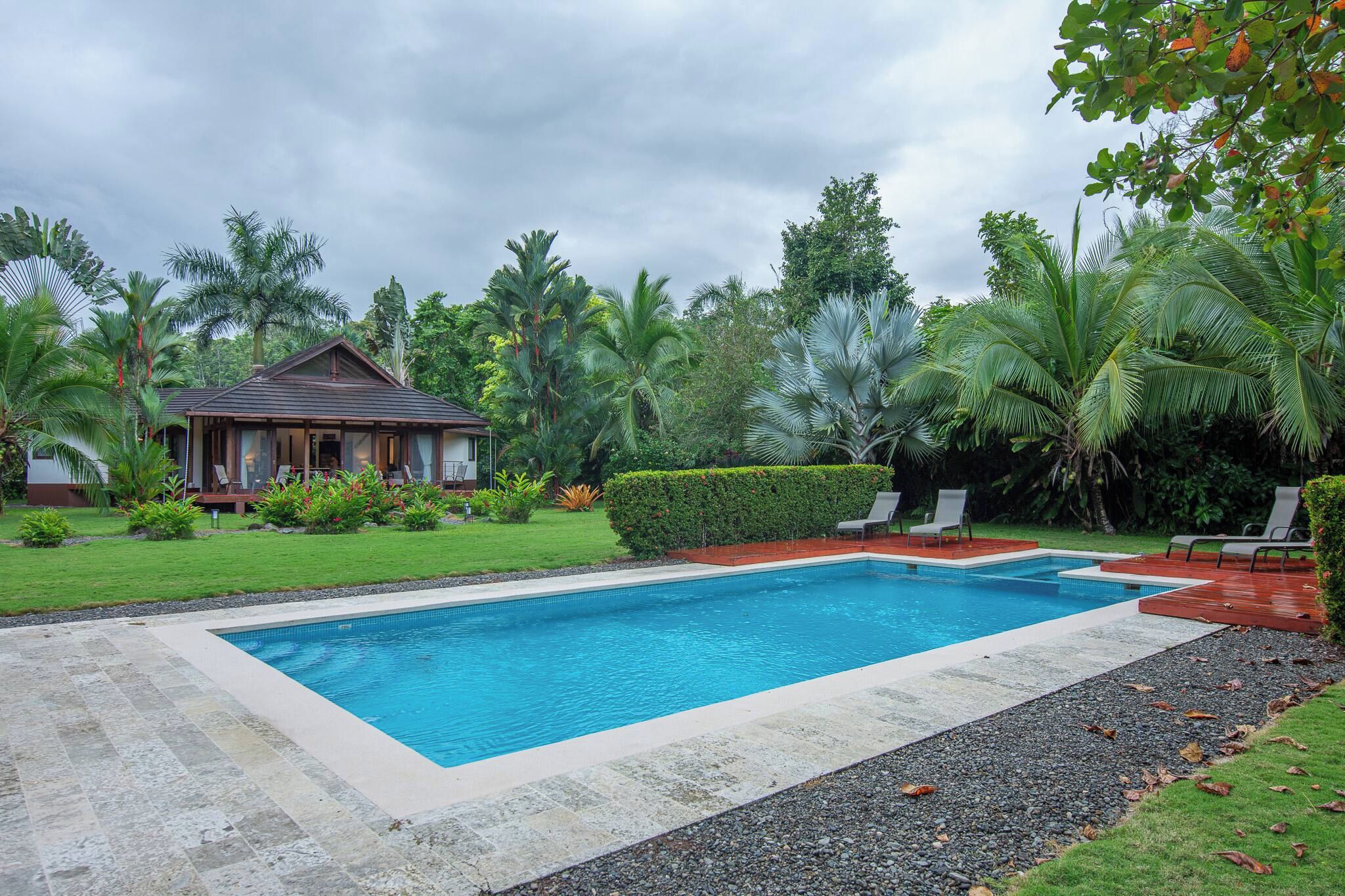 A rectangular swimming pool with a wooden deck surrounding it, a small house with a porch, and lush tropical vegetation.