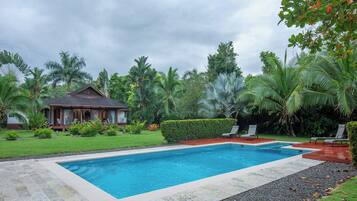 A rectangular swimming pool with a wooden deck surrounding it, a small house with a porch, and lush tropical vegetation.