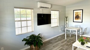 A modern living room with a flat-screen TV, a dining area with a white table and chairs, and a large potted plant.