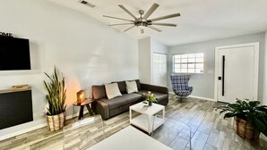 A modern living room with a grey sofa, a coffee table, and a TV mounted on the wall. There is a ceiling fan, a window with blinds, and a door.