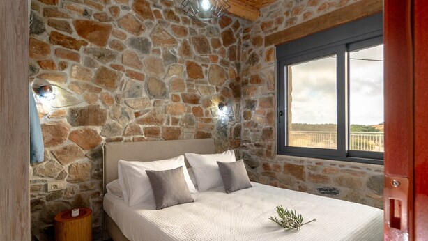 A bedroom with a stone wall, a bed with white bedding, a small wooden side table, and a window with a view of the landscape.