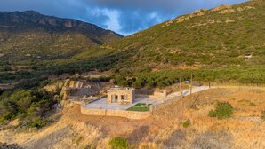A stone building with a small patio and a fenced area, surrounded by dry grass and sparse trees, with mountains in the background.