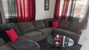 Ein Wohnzimmer mit einem Ecksofa, einem Glastisch und roten Vorhängen.