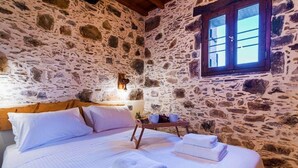 A stone-walled room with a bed, pillows, a tray with towels, and a window.