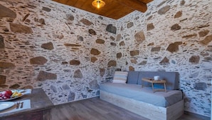 A cozy room with a stone wall, a wooden bed, a small table with a cup, and a fruit bowl on a wooden table.