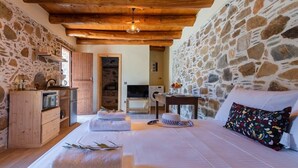 A cozy bedroom with a stone wall, wooden ceiling beams, a bed with white linens, a small table, and a stone fireplace. - Cozy Studio Plagia Room 3 (Plagia)