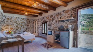 A cozy room with a stone wall, wooden ceiling, a bed, a small kitchen area, and a view of the countryside. - Cozy Studio Plagia Room 3 (Plagia)