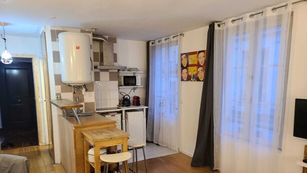 Et kompakt køkken med mikroovn, komfur og vask, et lille træbord med stole, og en seng i det tilstødende værelse.