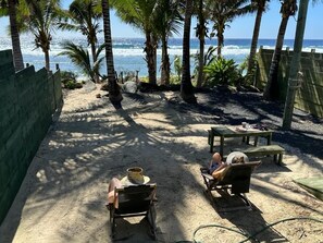 Ein Strand mit Palmen und einem Holzzaun. Zwei Holzstühle und ein Tisch, an dem eine Person sitzt.