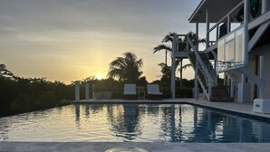 Una piscina con un solarium in legno, sedie a sdraio e una scalinata che porta a un edificio con un balcone.