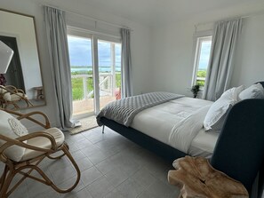 A bedroom with a bed, a chair, a wooden side table, and a balcony with a view.