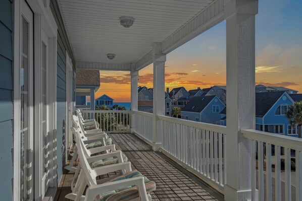 A covered porch with white railings and a view of houses across the water during sunset.