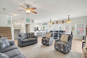 A spacious living room with a gray sofa, patterned armchairs, a ceiling fan, and a dining area with a kitchen in the background.