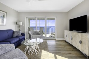 A living room with a blue sofa, a glass coffee table, a grey armchair, and a television on a cabinet. - Bayside Villa 5344- Stunning Direct Bay Views- Heated Pool (Captiva)