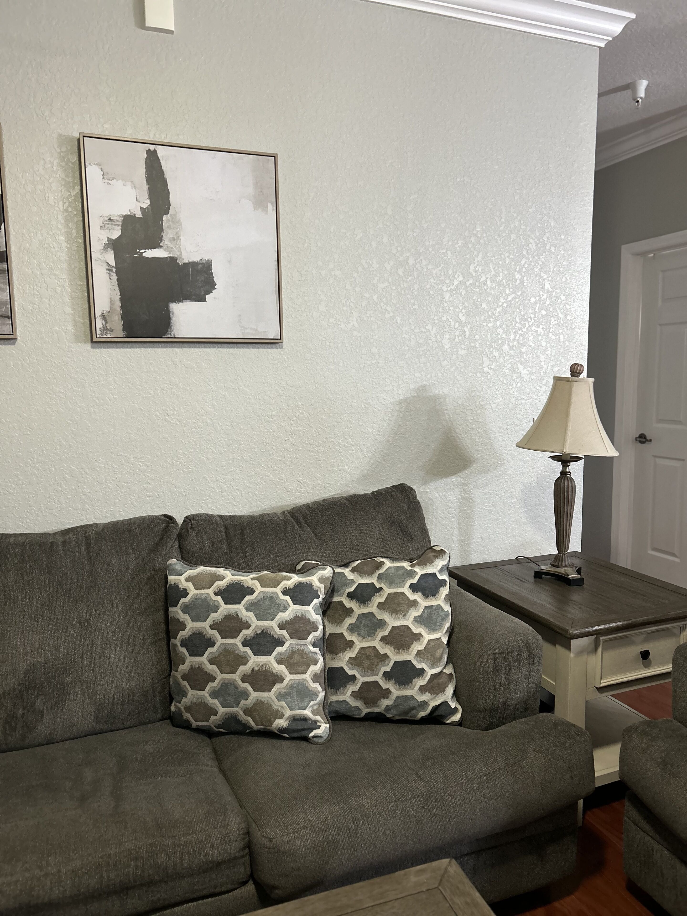 Een woonkamer met een grijze bank, gekleurde kussens, een bijzettafeltje met een lamp, en een lijst met een abstract schilderij aan de muur.