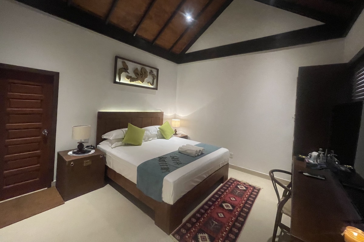 A bedroom with a wooden ceiling, a bed with a white mattress and green pillows, a bedside table with a lamp, and a patterned rug.