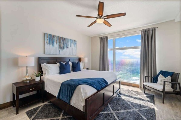 Une chambre d’hôtel avec un grand lit, un ventilateur de plafond, une fenêtre avec des rideaux, une table de chevet avec une lampe et une chaise.