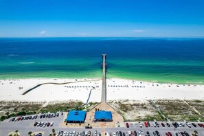 A beach with a pier extending into the ocean, a parking lot, and a building with a blue roof.