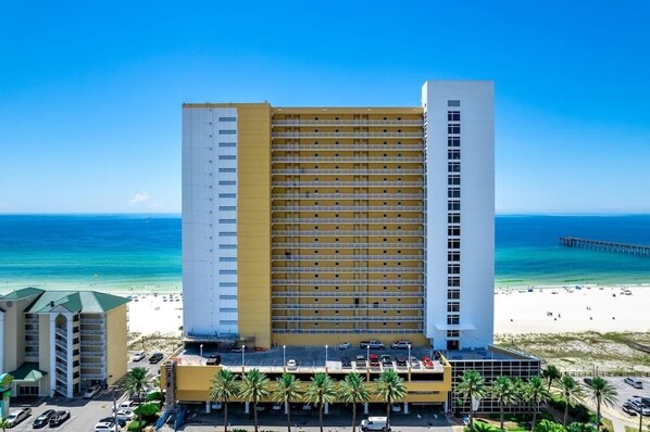 A multi-story hotel with a beach view, a parking lot, and a pier in the background.