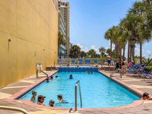 A swimming pool with people in the water, surrounded by palm trees and a building.