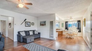 Un vaste espace de vie doté d’un canapé gris, de parquet, de ventilateurs de plafond et d’un coin repas avec des chaises blanches et une table.