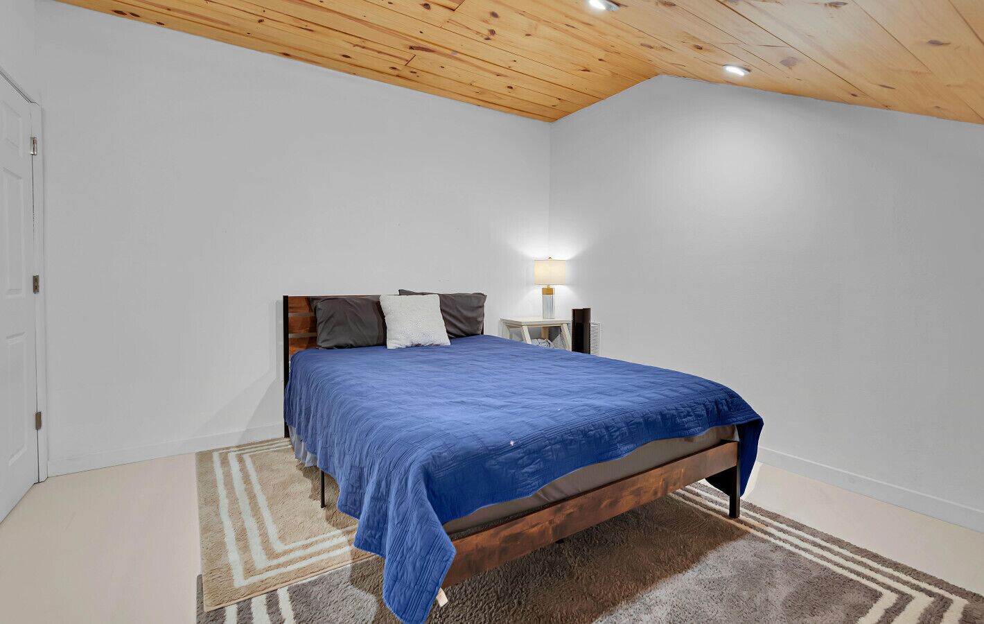 A bedroom with a wooden ceiling, a bed with a blue bedspread, a nightstand with a lamp, and a patterned rug.