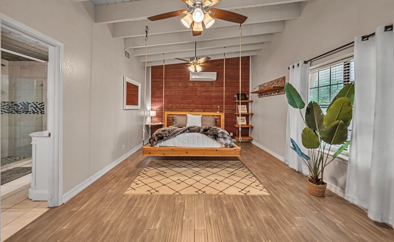 Une chambre avec un lit en bois, un ventilateur de plafond, une grande fenêtre avec des rideaux et une plante en pot.