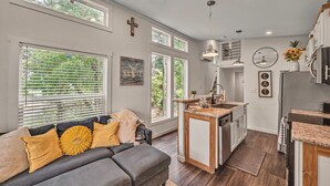 Una cocina moderna con una isla central, un área de comedor con un sofá gris y ventanas amplias con persianas.