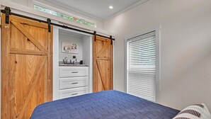 Un dormitorio con cama, armario con puerta corredera tipo granero, ventana con persianas y una pequeña zona de cocina con fregadero y armarios.