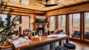 Interior de una cabaña de madera con chimenea, una zona de bar y amplias ventanas que ofrecen vistas al exterior.