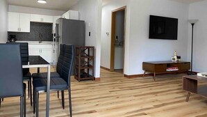 Un salon moderne avec un plancher en bois, un coin repas avec des chaises noires, une cuisine avec des meubles blancs et un téléviseur fixé au mur.