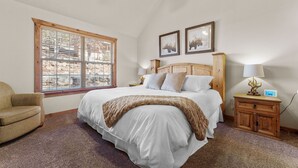 A bedroom with a large bed, a wooden headboard, a window with blinds, a bedside table with a lamp, and a chair.