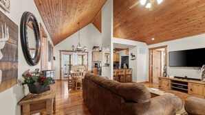 A spacious living room with a large brown leather sofa, wooden flooring, a wooden ceiling, and a flat-screen TV mounted on the wall.