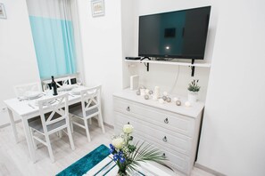 A modern, well-lit room with a flat-screen TV mounted on the wall, a white dining table set for two, and a white dresser with decorative items.