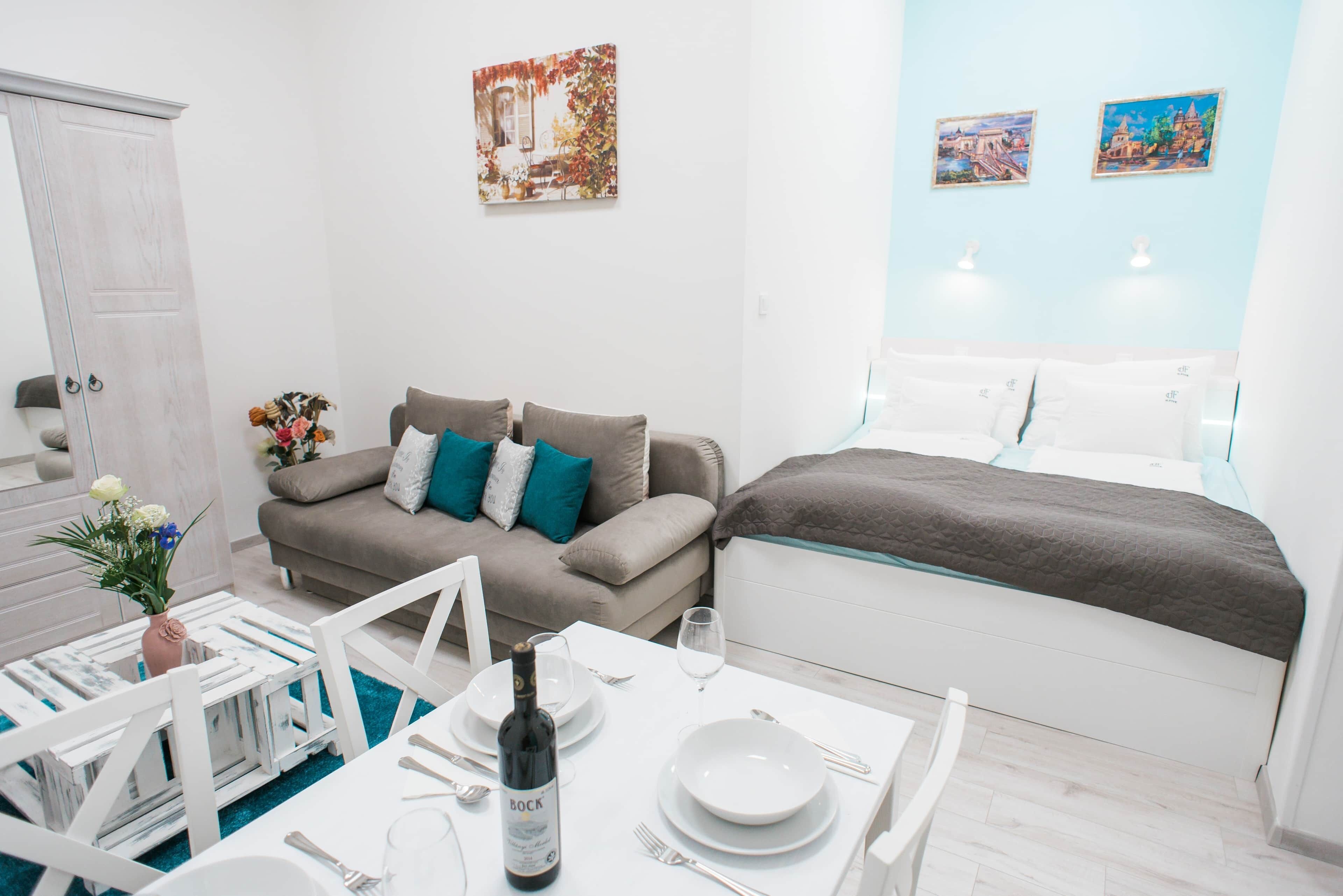 A modern, well-lit room with a sofa, a dining table set for two, and a bed with a quilt.