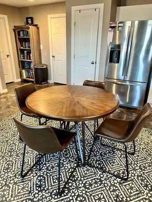 A dining area with a round wooden table, four chairs, a patterned rug, a bookshelf, and a stainless steel refrigerator.