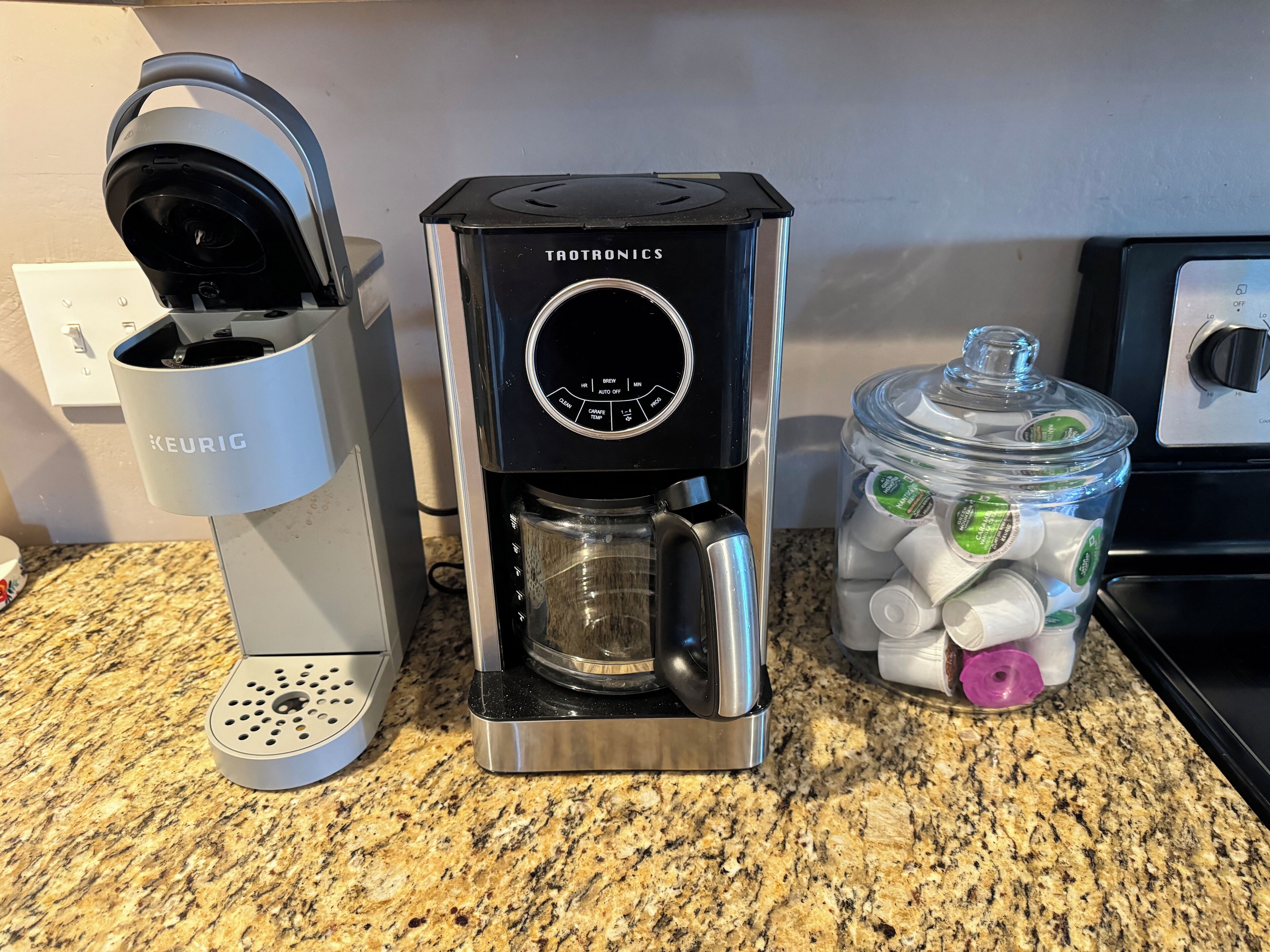 En Keurig-kaffemaskin med en Trovtronic-dispenser för kaffekapslar, ett Keurig-stativ för kaffekapslar och en behållare med kaffekapslar placerad på en köksbänk.