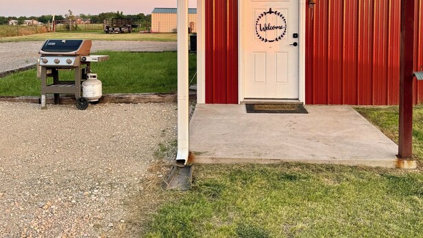 Un bâtiment rouge avec une porte blanche, un barbecue au gaz et un chemin de gravier.