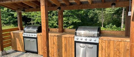 Una estación de parrilla al aire libre con dos asadores a gas y un horno empotrado, frente a un fondo de madera y suelo de grava.