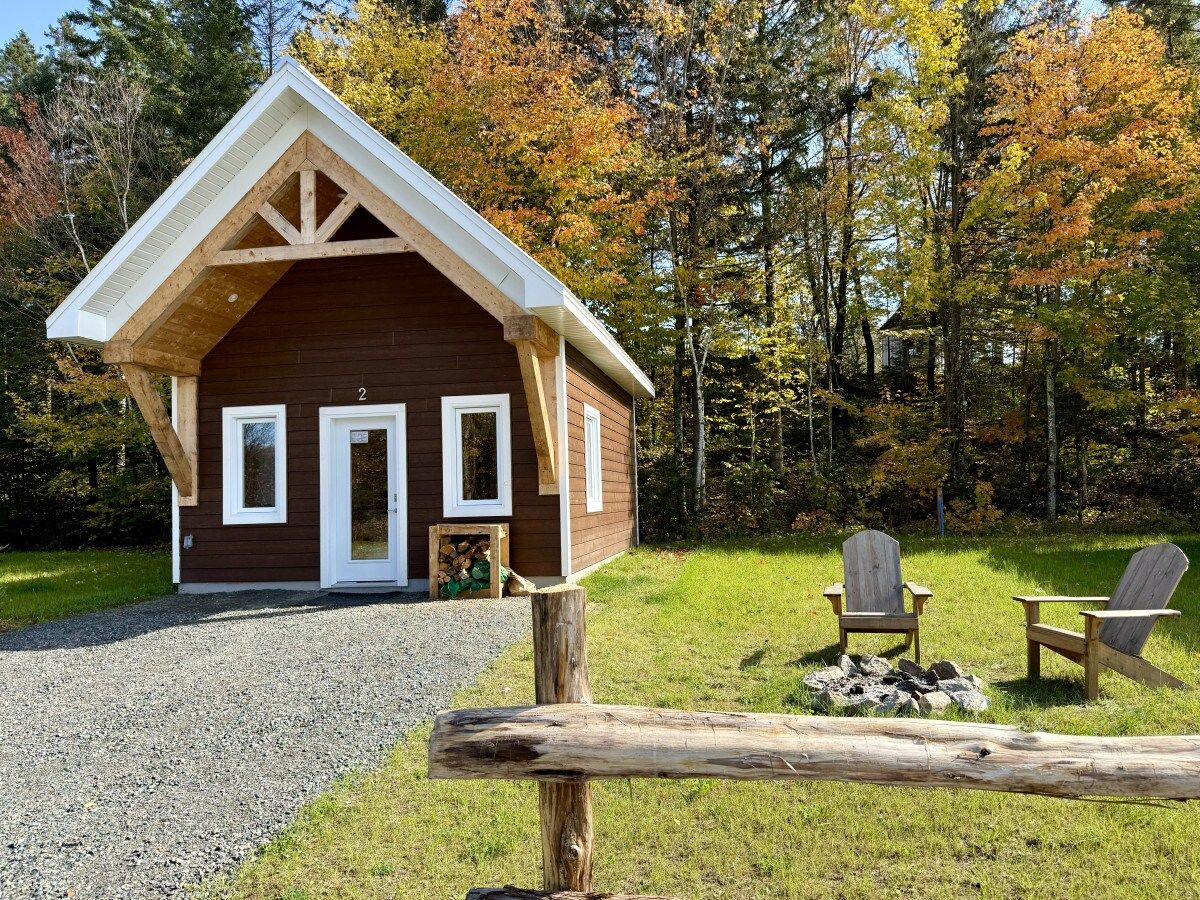 A cabin with a pitched roof, a gravel driveway, and two Adirondack chairs in front.