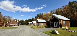 A row of cabins with a gravel driveway, surrounded by trees and a clear sky. - Mini-Chalet #2 – Near the River (Saint-Gabriel-de-Valcartier)