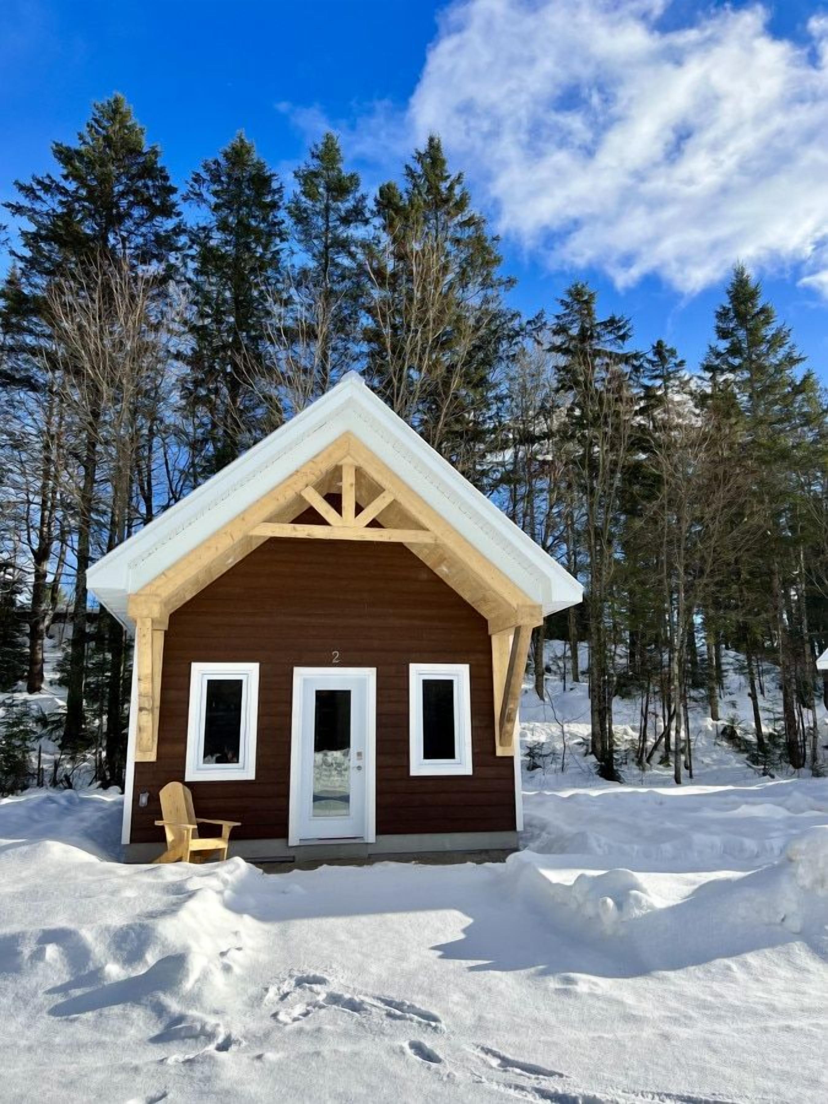 A small wooden cabin with a gabled roof, a door, and two windows, surrounded by snow and trees.